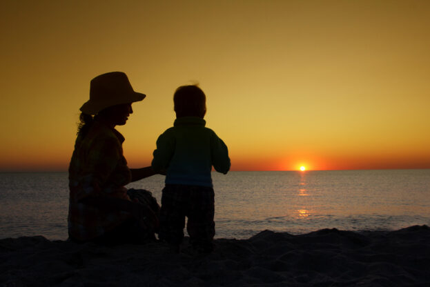 Mother with little baby sitting together on sunset beach. Family vacation on the sea.